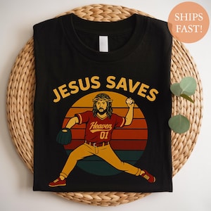 May include: Black t-shirt with a graphic of Jesus as a baseball pitcher. The design includes the text "JESUS SAVES" in yellow, and Jesus is wearing a red baseball jersey with "Heaven 01". The background is a sunset.