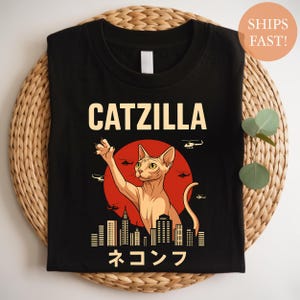 May include: Black t-shirt featuring a cartoon cat with the text "CATZILLA" above and Japanese text below. The cat is depicted in front of a red sun with a city skyline and helicopters.