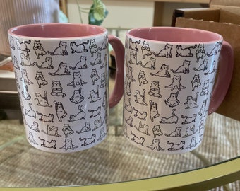 May include: Two white ceramic mugs with pink rims and handles. The mugs are covered in a pattern of black line drawings of cats doing yoga poses.