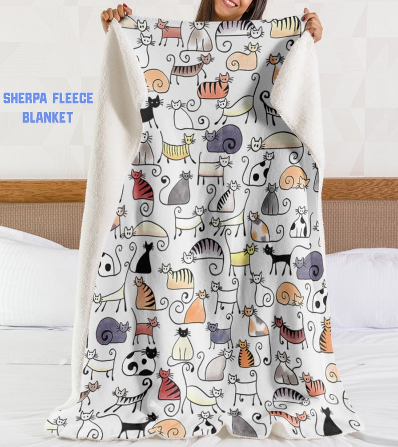 May include: A white sherpa fleece blanket with a colorful cat print. The cats are drawn in a simple, cartoon style and are all different colors and patterns. The blanket is soft and cozy and would be perfect for cuddling up on the couch.
