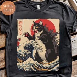 May include: Black t-shirt featuring a stylized illustration of a black and white cat with a red sun in the background, inspired by Japanese art. The cat is positioned on a wave, with Japanese text. The shirt has a round neck.