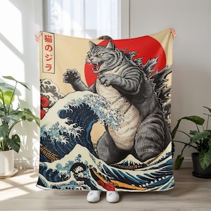 May include: A plush blanket featuring a gray cat in a Godzilla-like pose, set against a red sun and a stylized ocean wave design. The blanket has Japanese text on the side and is held up by a person.