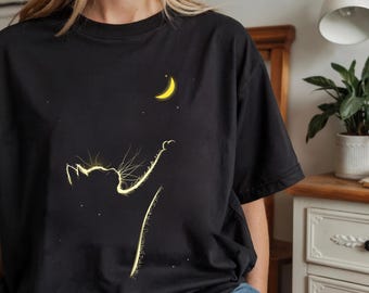 Op de afbeelding: Zwart T-shirt met een geel silhouet van een kat die naar een gele halve maan reikt met sterren op de achtergrond.