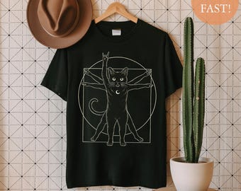 Puede incluir: Camiseta negra con un diseño blanco de un gato en la pose del Hombre de Vitruvio, con una luna creciente y un gesto de la mano de rock. Un sombrero marrón cuelga a la izquierda. El diseño está sobre un fondo negro.