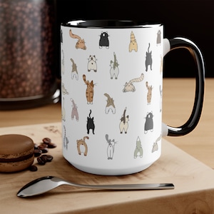 May include: White ceramic mug with a black rim and handle. The mug features a repeating pattern of cartoon cats with their tails up in the air.