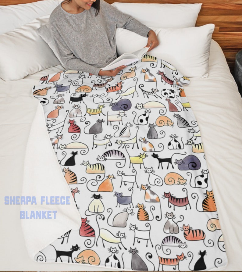 May include: White sherpa fleece blanket with a colorful cat print. The blanket features a repeating pattern of stylized cats in various colors and poses. The text "Sherpa Fleece Blanket" is printed in the bottom left corner.
