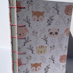 May include: A white fabric-covered journal with a pink stitching detail. The cover features a repeating pattern of animal faces, including foxes, bears, pandas, and cats, with small floral sprigs in between.