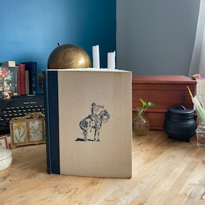 May include: A vintage hardcover book with a blue spine and a beige cover. The cover features a black and white illustration of a person on horseback blowing a trumpet.