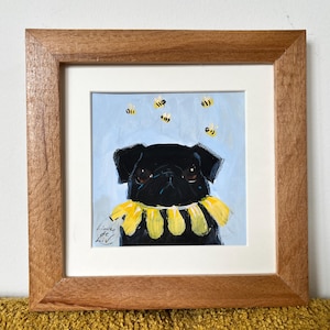 May include: A framed painting of a black pug wearing a yellow petal collar against a light blue background with small yellow bees. The artwork is signed Lieve de Liv in the bottom left corner and encased in a square wooden frame.