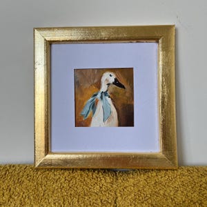 May include: A square painting of a white duck wearing a light blue ribbon around its neck, set against a warm brown background. The artwork is encased in a thick, textured gold frame with a white mat border surrounding the central painted image.