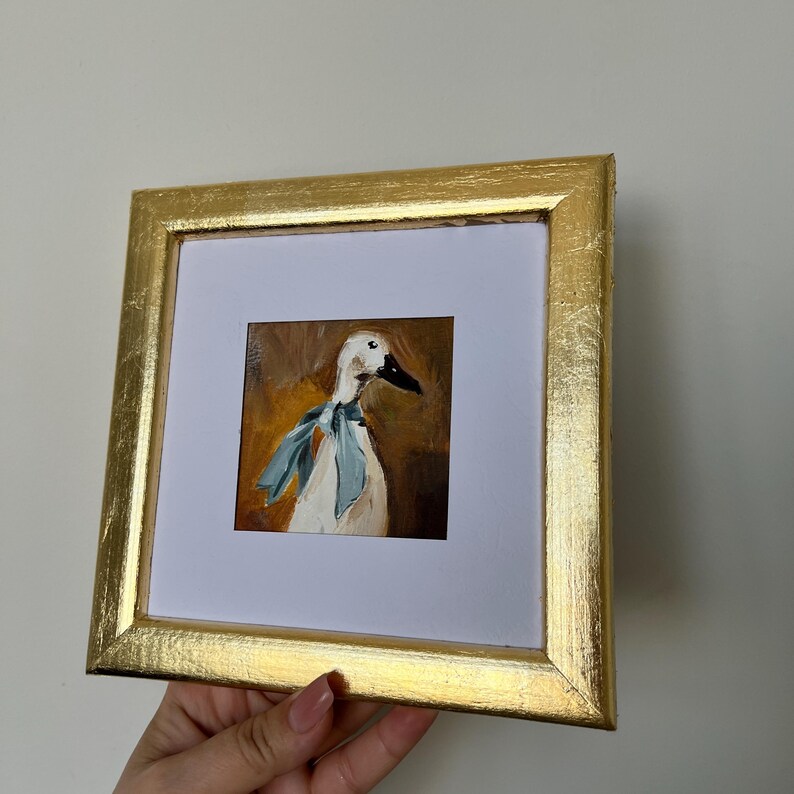 May include: A square framed painting featuring an oil-style portrait of a white duck wearing a light blue neck ribbon. The artwork is set against a warm brown background and is encased in a textured, metallic gold frame with a white mat border.