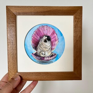Op de afbeelding: Een vierkante houten lijst met een cirkelvormig schilderij van een mopshond die in een grote, open roze schelp zit. De achtergrond van het schilderij is lichtblauw en de mopshond is afgebeeld met zijn kenmerkende gerimpelde gezicht en donkere oren.