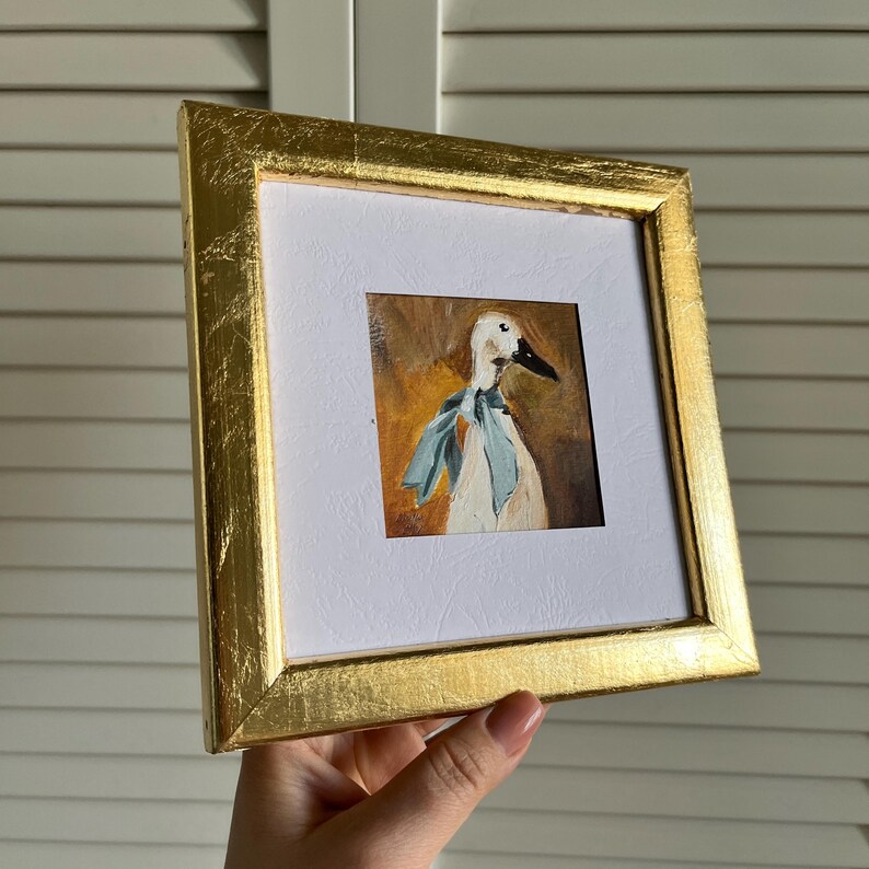 May include: A square painting of a white duck wearing a light blue ribbon around its neck, set against a warm brown background. The artwork is encased in a textured, metallic gold frame with a white mat border surrounding the central image.