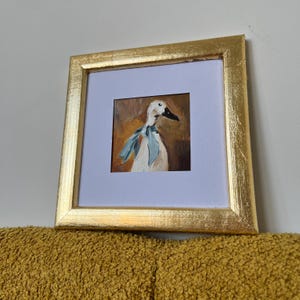 May include: A square painting of a white duck wearing a light blue ribbon around its neck, set against a warm brown background. The artwork is enclosed in a textured gold frame with a white mat border, presenting a clear view of the painted subject.
