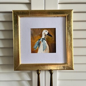May include: A square painting of a white duck wearing a light blue neck ribbon, set against a warm brown background. The artwork is encased in a textured white mat and a reflective gold frame with a visible metallic finish and square corners.