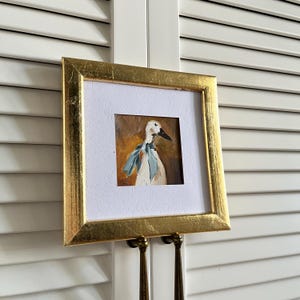 May include: A square painting of a white duck wearing a light blue neck scarf, set against a warm brown background. The artwork is enclosed in a textured gold frame with a white mat border, creating a distinct contrast between the frame and art.