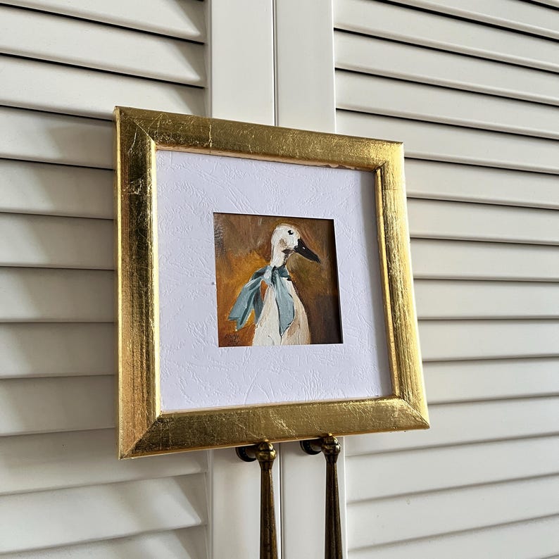 May include: A square painting of a white duck wearing a light blue neck scarf, set against a warm brown background. The artwork is enclosed in a textured gold frame with a white mat border, creating a distinct contrast between the frame and art.