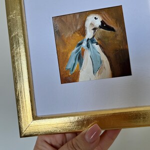 May include: A square painting of a white duck wearing a light blue neck scarf, rendered in visible brushstrokes against a warm brown background. The artwork is encased in a metallic gold frame with a white mat border surrounding the central image.