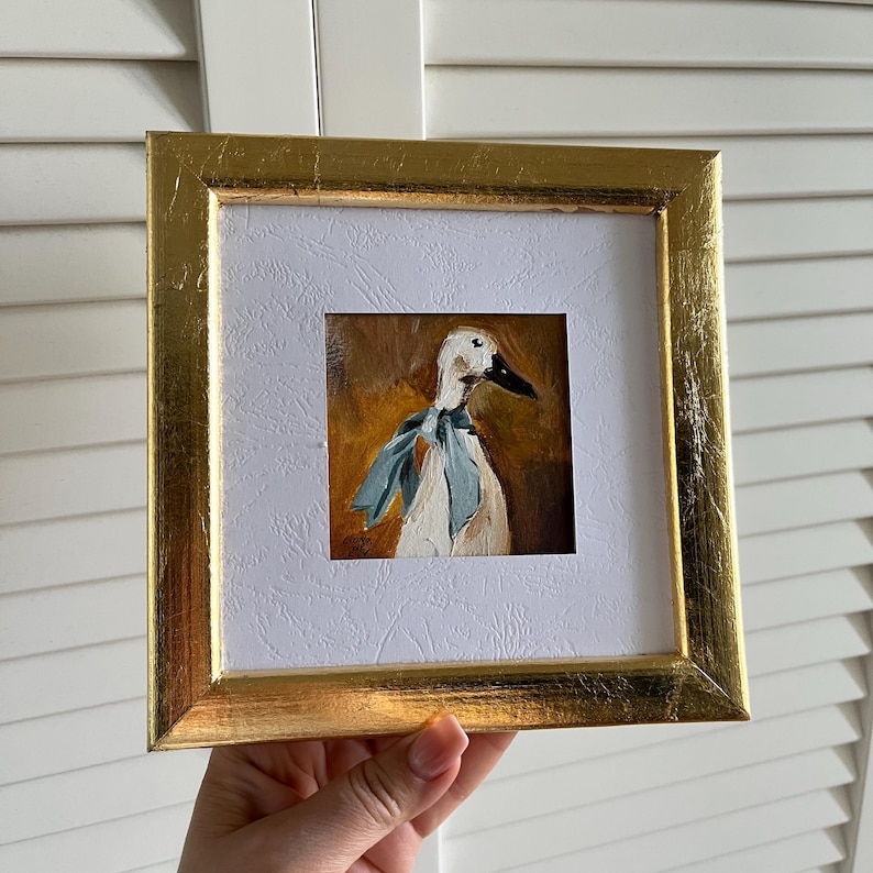 May include: A square oil painting of a white duck wearing a light blue neck ribbon, set against a warm brown background. The artwork is encased in a bright gold frame with a textured white mat board surrounding the central painted image.