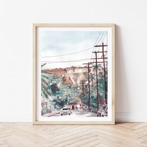 May include: A watercolour painting depicting a street scene in Los Angeles, California. The Hollywood sign is visible in the distance on a hillside. The street is lined with palm trees and power lines. There are cars driving on the street.