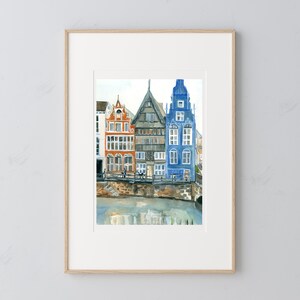 May include: A framed watercolor painting depicting three narrow, multi-story buildings in orange, grey, and blue, situated along a stone embankment beside a body of water. The artwork features detailed architectural facades with multiple windows.