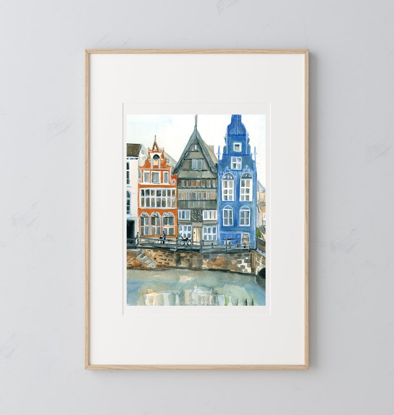 May include: A framed watercolor painting depicting three narrow, multi-story buildings in orange, grey, and blue, situated along a stone embankment beside a body of water. The artwork features detailed architectural facades with multiple windows.