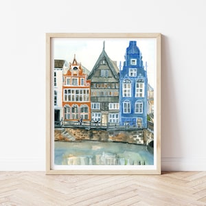 May include: A framed watercolor painting depicting a row of three narrow European-style buildings in orange, grey, and blue, situated along a stone embankment above a canal with reflections visible in the water below the structures.