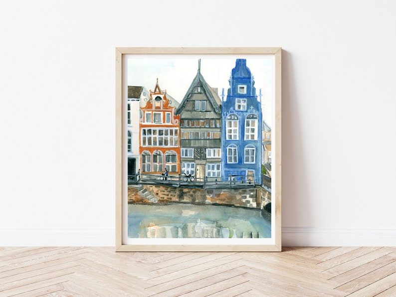 May include: A framed watercolor painting depicting a row of three narrow European-style buildings in orange, grey, and blue, situated along a stone embankment above a canal with reflections visible in the water below the structures.