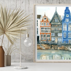 May include: A framed watercolor painting depicting a row of tall, narrow houses in orange, grey, and blue colors situated along a canal. The artwork features detailed architectural facades with multiple windows, reflecting in the water below the stone embankment.