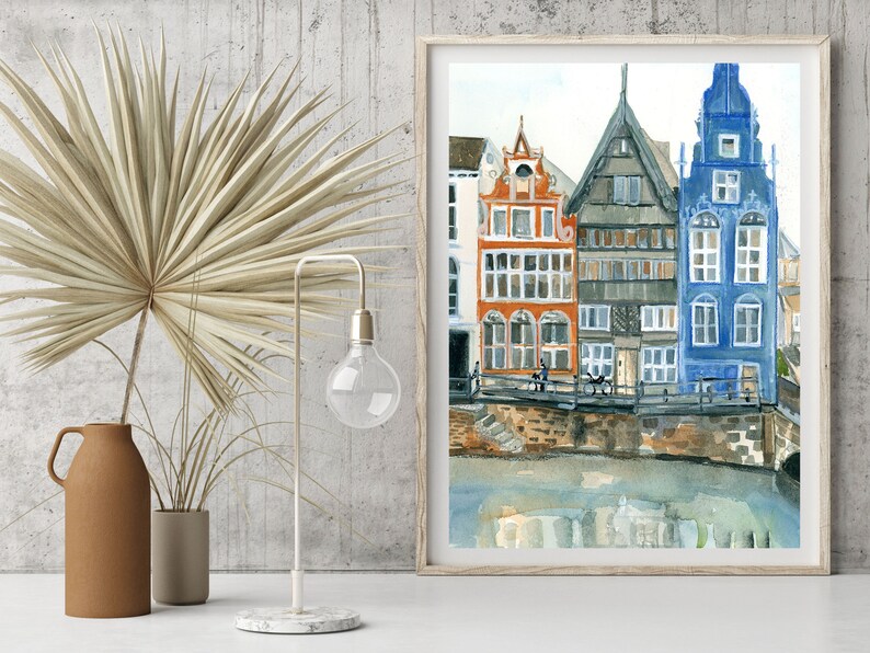 May include: A framed watercolor painting depicting a row of tall, narrow houses in orange, grey, and blue colors situated along a canal. The artwork features detailed architectural facades with multiple windows, reflecting in the water below the stone embankment.