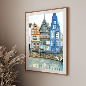 May include: A framed watercolor painting depicting a row of three narrow, multi-story buildings in orange, gray, and blue, situated along a stone embankment above a body of water, with architectural details including multiple windows and gables.