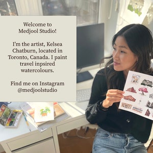 May include: A woman sits at a desk with a white surface. She is holding a sheet of paper with watercolor illustrations of buildings. The paper has the text "Welcome to Medjool Studio! I'm the artist, Kelsea Chatburn, located in Toronto, Canada. I paint travel inspired watercolours. Find me on Instagram @medjoolstudio".