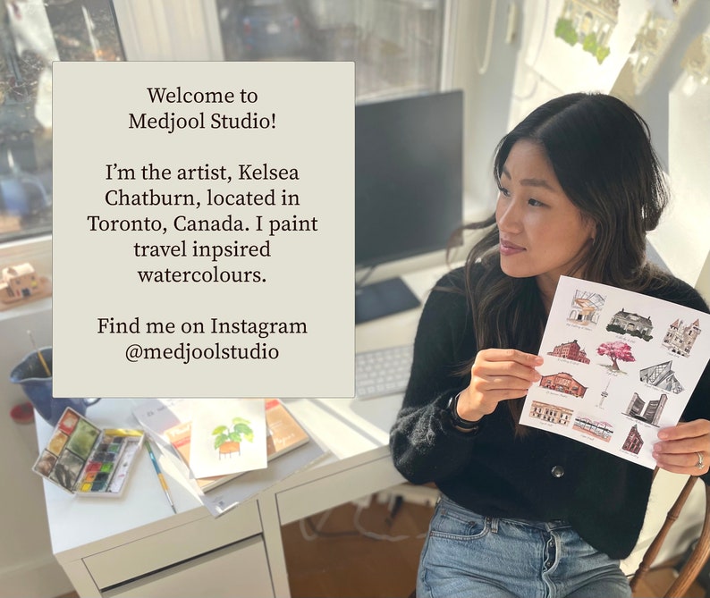 May include: A woman sits at a desk with a white surface. She is holding a sheet of paper with watercolor illustrations of buildings. The paper has the text "Welcome to Medjool Studio! I'm the artist, Kelsea Chatburn, located in Toronto, Canada. I paint travel inspired watercolours. Find me on Instagram @medjoolstudio".