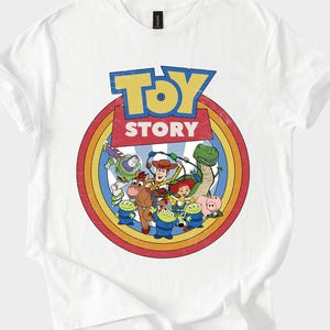 Puede incluir: Camiseta blanca con un dise&ntilde;o gr&aacute;fico colorido de los personajes de la pel&iacute;cula "Toy Story". El dise&ntilde;o incluye el logotipo de la pel&iacute;cula en azul y amarillo, rodeado por un c&iacute;rculo de colores del arco&iacute;ris e ilustraciones de personajes.
