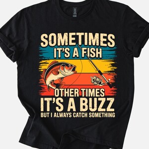 May include: Black t-shirt with a fishing-themed graphic. The design features a red fish, a fishing rod, and the text "SOMETIMES IT'S A FISH, OTHER TIMES IT'S A BUZZ, BUT I ALWAYS CATCH SOMETHING" in a colorful, retro style.