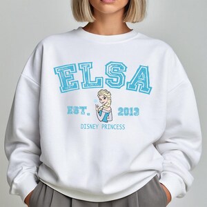 K&ouml;nnte beinhalten: Wei&szlig;es Sweatshirt mit dem Namen "ELSA" in hellblauen Blockbuchstaben. Darunter steht "EST. 2013" und eine Grafik von Elsa aus Disneys Frozen. Die Worte "DISNEY PRINCESS" sind darunter gedruckt.