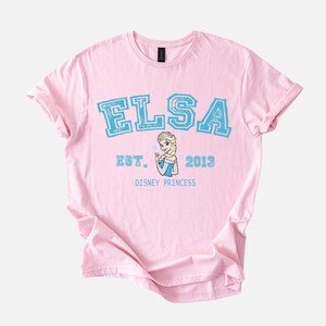 K&ouml;nnte beinhalten: Ein hellrosa T-Shirt mit dem Namen "ELSA" in blauen Blockbuchstaben. Darunter steht "EST. 2013" und eine Grafik von Elsa aus Disneys Frozen. Der Text "DISNEY PRINCESS" befindet sich darunter.