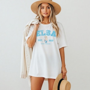 K&ouml;nnte beinhalten: Wei&szlig;es T-Shirt mit dem Namen "ELSA" in hellblauen Blockbuchstaben. Das Shirt hat auch den Text "EST. 2013" und eine Grafik einer Disney-Prinzessin. Das Model tr&auml;gt einen Strohhut und h&auml;lt eine Jacke.