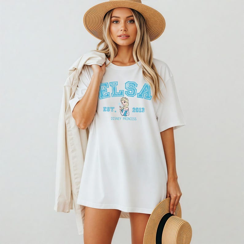 K&ouml;nnte beinhalten: Wei&szlig;es T-Shirt mit dem Namen "ELSA" in hellblauen Blockbuchstaben. Das Shirt hat auch den Text "EST. 2013" und eine Grafik einer Disney-Prinzessin. Das Model tr&auml;gt einen Strohhut und h&auml;lt eine Jacke.