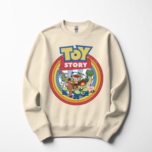 Puede incluir: Sudadera de color crema con un gr&aacute;fico colorido de personajes de la pel&iacute;cula animada "Toy Story". El dise&ntilde;o incluye las palabras "Toy Story" en una fuente azul y amarilla, rodeadas por un c&iacute;rculo de arco&iacute;ris.