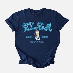 K&ouml;nnte beinhalten: Marineblaues T-Shirt mit dem Namen "ELSA" in hellblauen Blockbuchstaben und "EST. 2013" darunter. Ein Cartoon-Bild von Elsa, die eine Schneeflocke h&auml;lt, ist unter dem Text zentriert. Die Worte "DISNEY PRINCESS" sind unter dem Bild gedruckt.