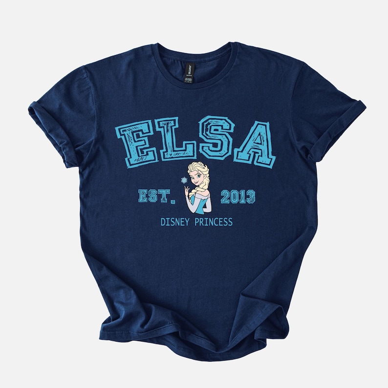 K&ouml;nnte beinhalten: Marineblaues T-Shirt mit dem Namen "ELSA" in hellblauen Blockbuchstaben und "EST. 2013" darunter. Ein Cartoon-Bild von Elsa, die eine Schneeflocke h&auml;lt, ist unter dem Text zentriert. Die Worte "DISNEY PRINCESS" sind unter dem Bild gedruckt.