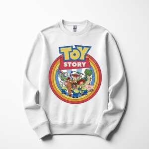 Puede incluir: Sudadera blanca de cuello redondo con un gr&aacute;fico colorido de personajes de la pel&iacute;cula animada "Toy Story". El dise&ntilde;o incluye el logotipo de la pel&iacute;cula en azul y amarillo, rodeado por un c&iacute;rculo de colores del arco&iacute;ris e im&aacute;genes de Woody, Buzz Lightyear y otros personajes.