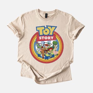 Puede incluir: Camiseta beige con un gr&aacute;fico colorido de personajes de la pel&iacute;cula animada "Toy Story". El dise&ntilde;o incluye las palabras "Toy Story" en azul y amarillo, rodeadas por un c&iacute;rculo de colores del arco&iacute;ris. La camiseta es de material suave.