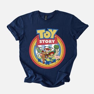 Puede incluir: Camiseta azul marino con un gr&aacute;fico colorido de la pel&iacute;cula "Toy Story". El dise&ntilde;o incluye el t&iacute;tulo de la pel&iacute;cula en amarillo y azul, rodeado de una imagen circular de los personajes. La camiseta est&aacute; hecha de un material suave.