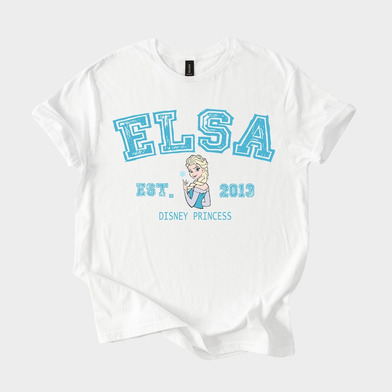 K&ouml;nnte beinhalten: Wei&szlig;es T-Shirt mit dem Namen "ELSA" in blauen Blockbuchstaben, "EST. 2013" und "DISNEY PRINCESS" darunter. Ein Cartoon-Bild von Elsa befindet sich in der Mitte.