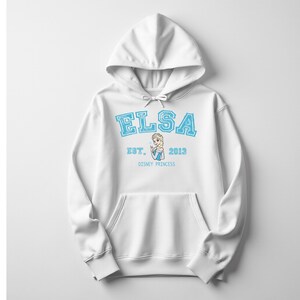 K&ouml;nnte beinhalten: Wei&szlig;er Kapuzenpullover mit dem Namen "ELSA" in hellblauen Blockbuchstaben. Darunter stehen die Worte "EST. 2013" und eine Grafik von Elsa, darunter "DISNEY PRINCESS". Der Pullover hat eine Vordertasche.