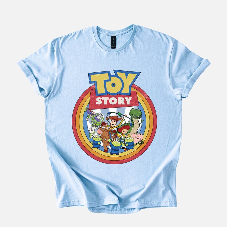Puede incluir: Camiseta azul claro con el logo de Toy Story en amarillo y rojo, con un dise&ntilde;o circular que muestra personajes de la pel&iacute;cula animada. La camiseta tiene mangas cortas y cuello redondo.