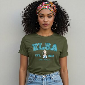 K&ouml;nnte beinhalten: Olivgr&uuml;nes T-Shirt mit dem Namen "ELSA" in blauen Blockbuchstaben, dem Text "EST. 2013" und einem Cartoon-Bild von Elsa. Das Shirt hat einen Rundhalsausschnitt mit hochgekrempelten &Auml;rmeln. Das Model tr&auml;gt ein buntes Kopftuch und Creolen.