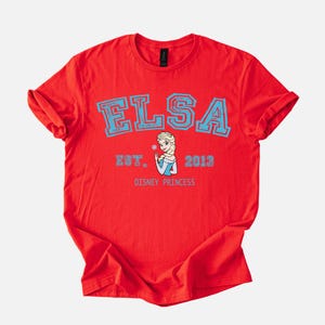 K&ouml;nnte beinhalten: Rotes T-Shirt mit dem Namen "ELSA" in hellblauen Blockbuchstaben. Darunter ein Bild von Elsa aus Disneys Frozen, mit dem Text "EST. 2013" und "DISNEY PRINCESS" in Hellblau.